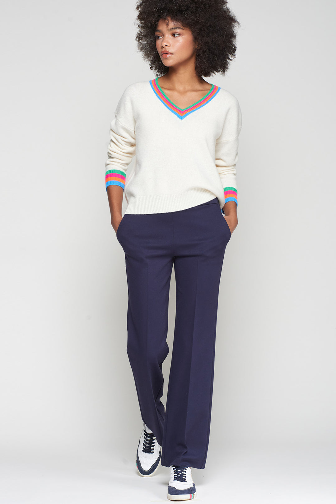 Woman wearing a white sweater with colorful trim and navy pants on a light gray background