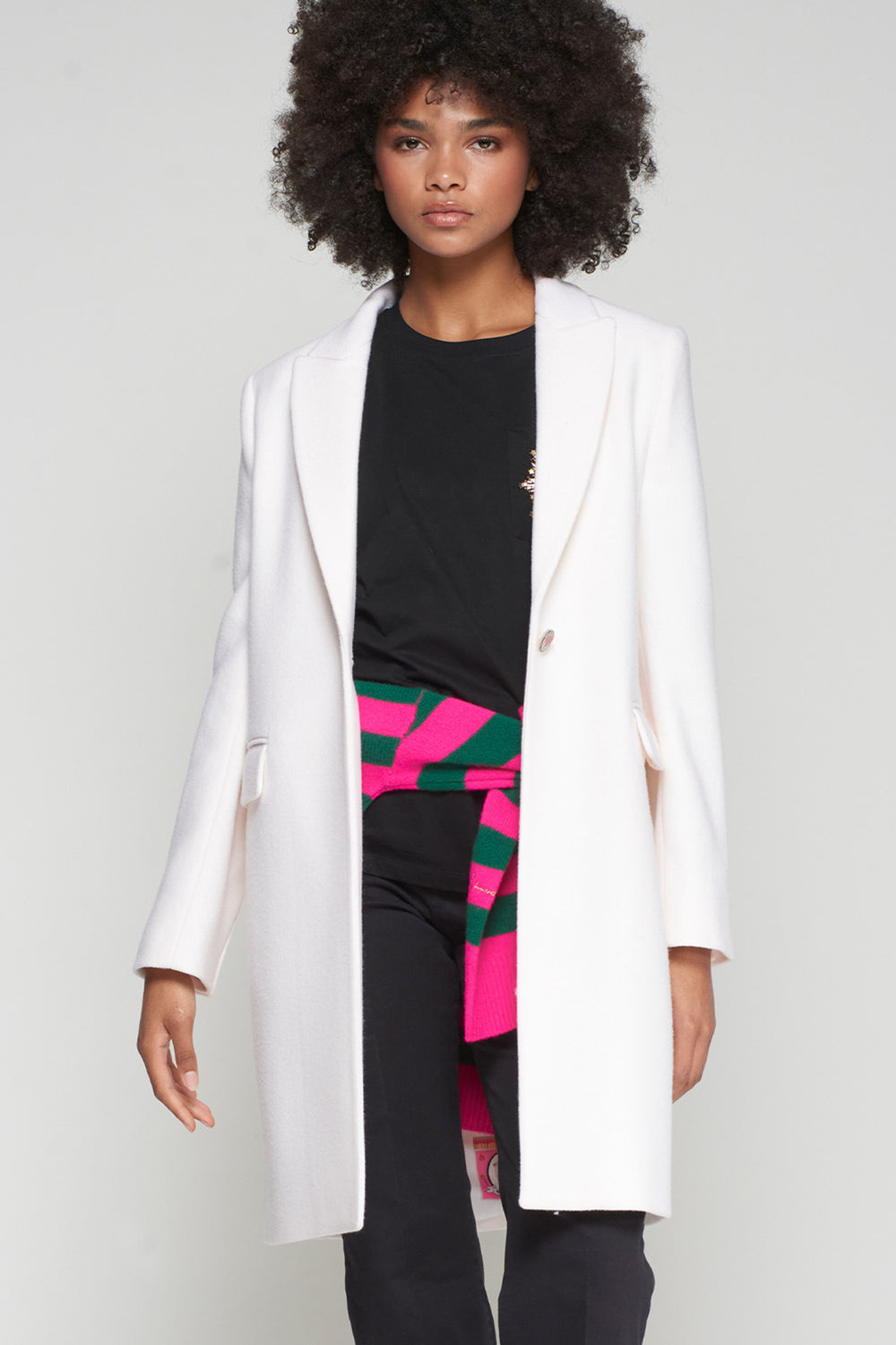 Person wearing a white coat over a black outfit with colorful pants on a plain background