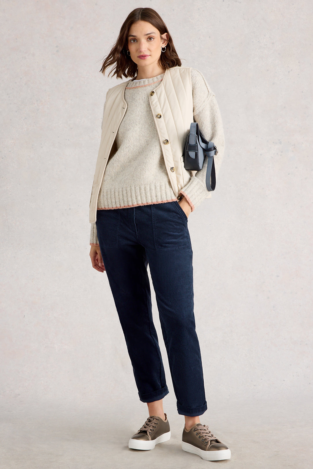 Woman wearing a beige cardigan, white sweater, navy pants, and brown shoes on a light gray background