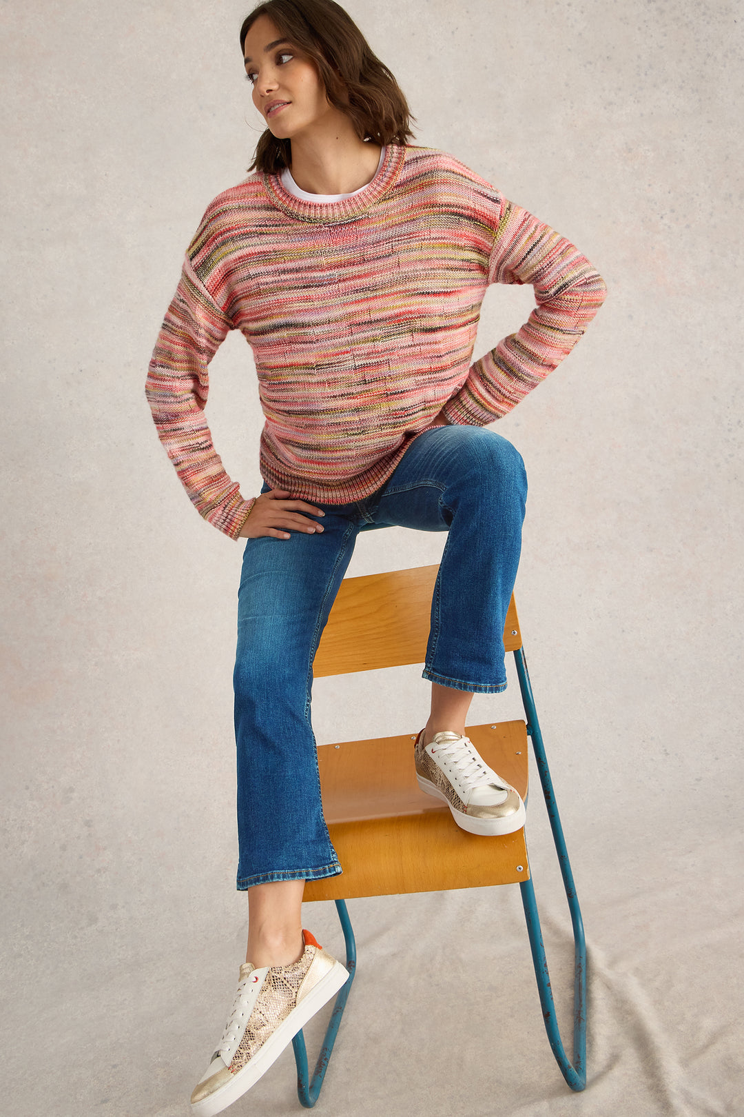 Woman wearing a colorful sweater and blue jeans sitting on a chair against a neutral background