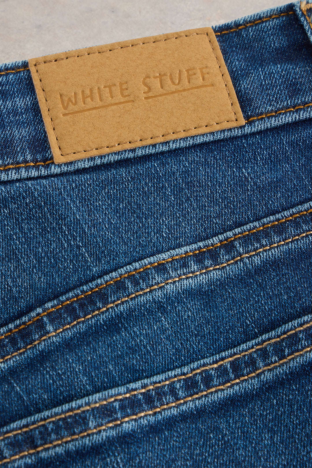 Close-up of blue denim jeans with a tan label reading 'White Stuff' on a neutral background.