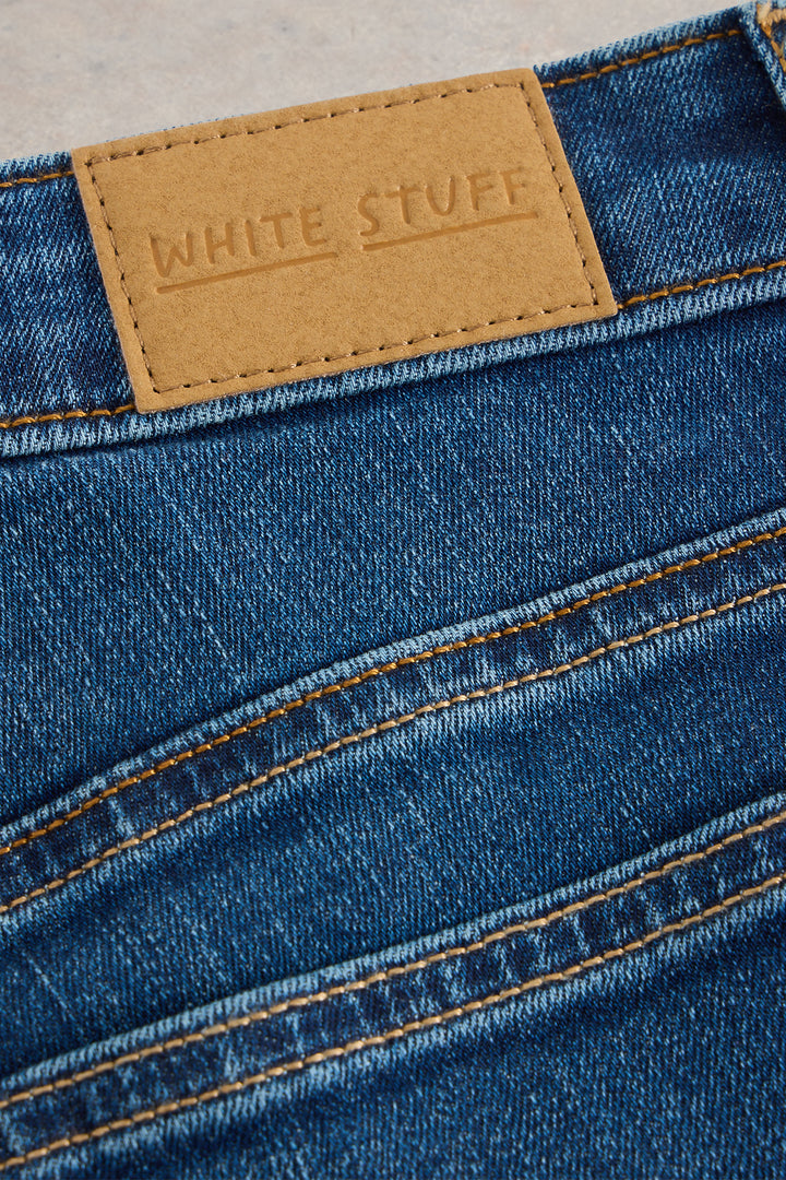 Close-up of blue denim jeans with a tan label reading 'White Stuff' on a neutral background.