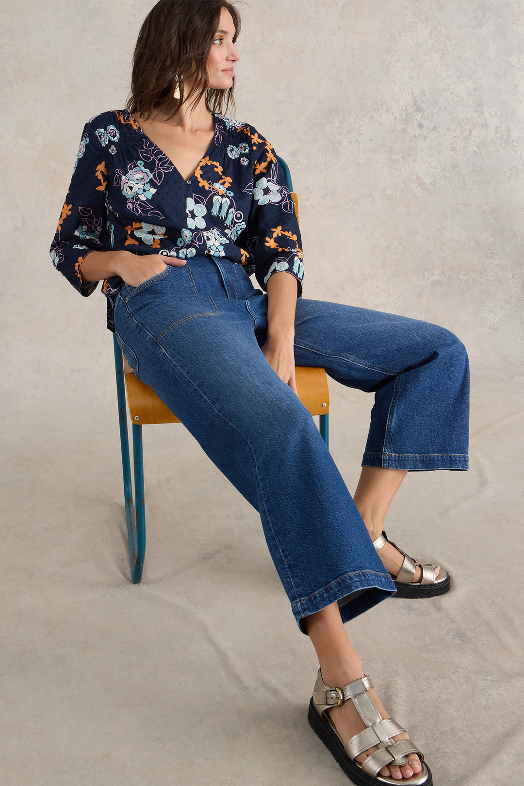 Woman sitting on a chair wearing a floral blouse and blue jeans against a neutral background