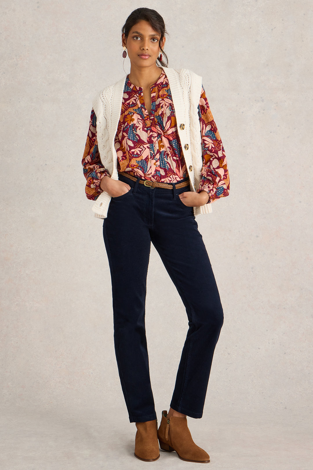 Woman wearing a floral blouse, white cardigan, dark jeans, and brown boots on a plain background