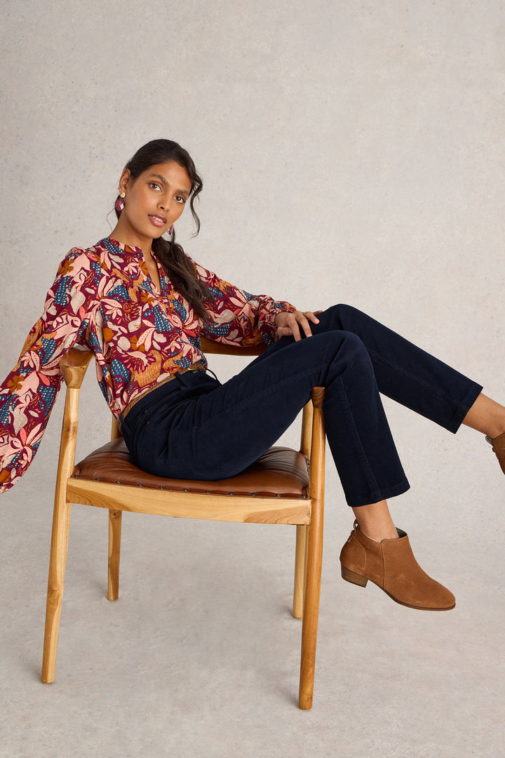 Woman sitting on a wooden chair wearing a patterned blouse and dark pants.