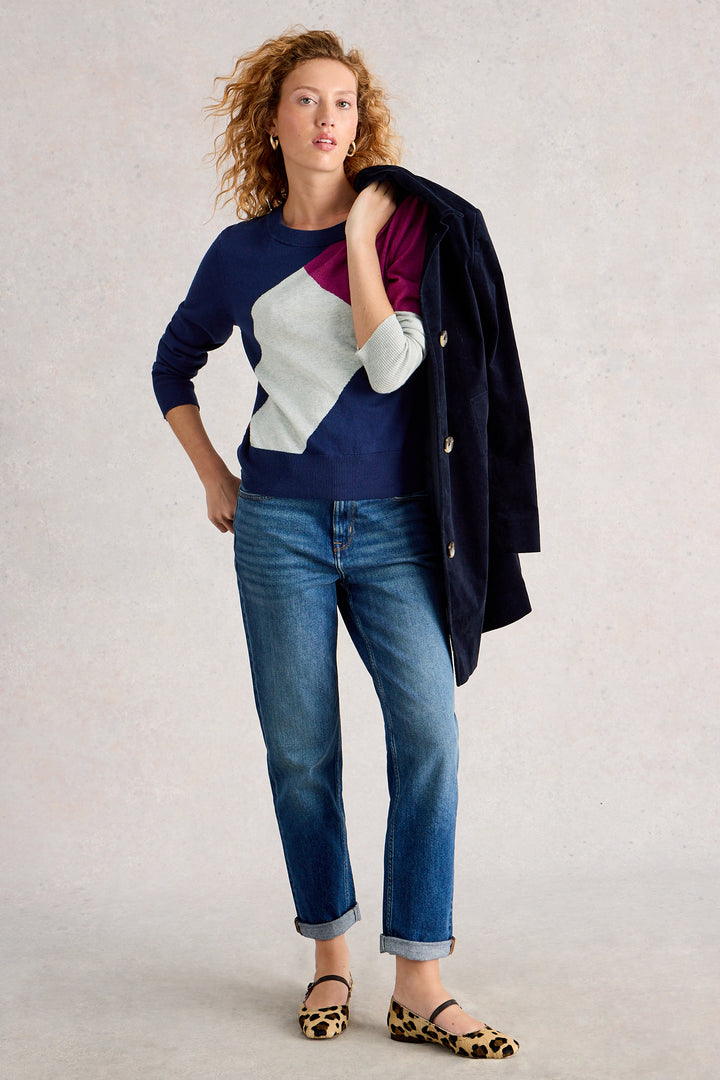 Woman wearing a colorful sweater and blue jeans with a black jacket over her shoulder on a light background