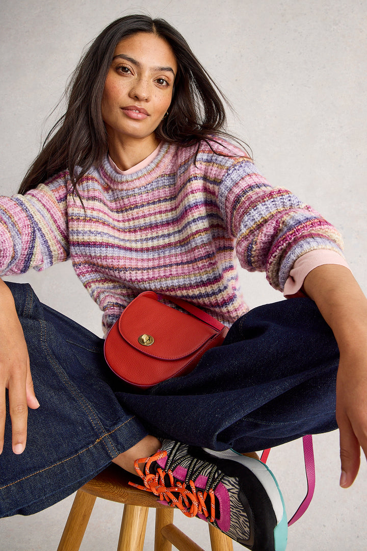 Woman wearing a colorful sweater and jeans, sitting on a stool with a red bag.