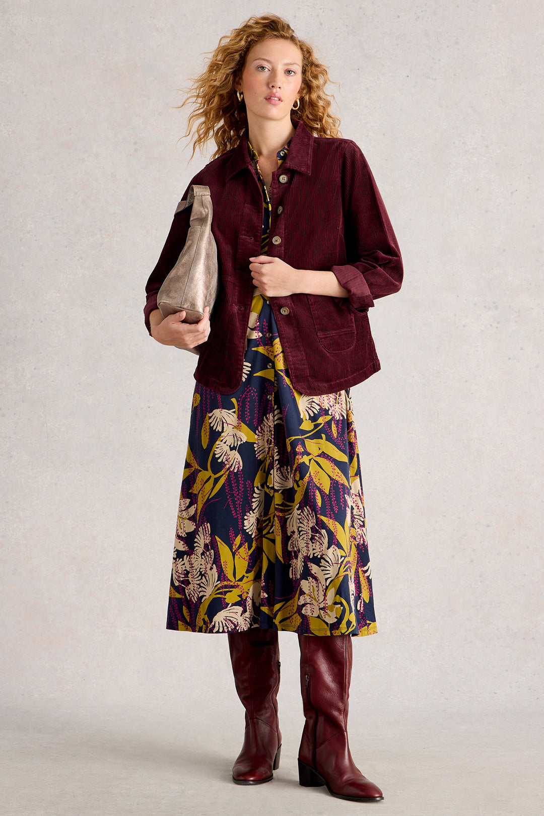 Woman wearing a burgundy jacket, floral dress, and brown boots against a plain background