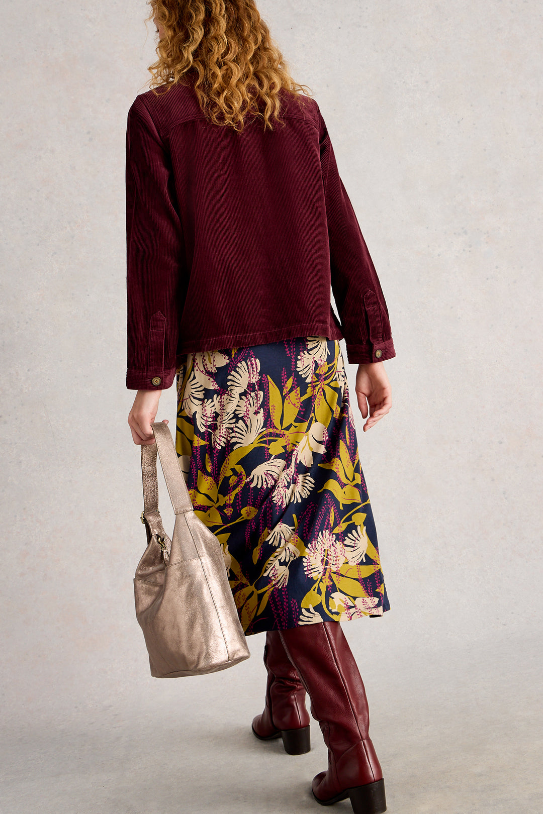 Person wearing a burgundy jacket, floral skirt, and red boots, holding a beige handbag against a plain background.