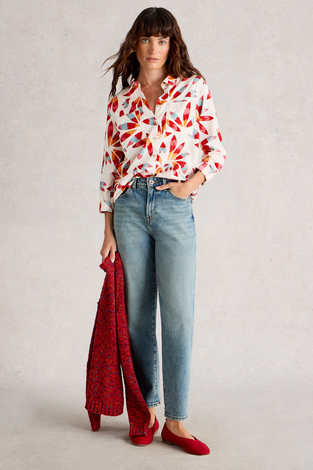 Woman wearing a colorful blouse, jeans, and holding a red jacket against a neutral background