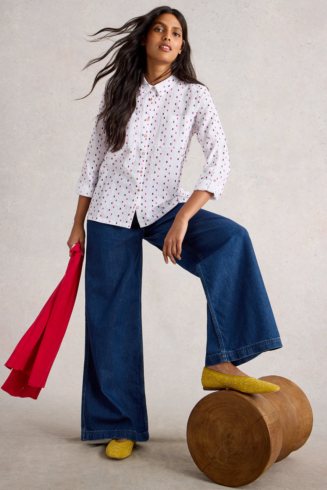 Woman in a white patterned shirt and blue jeans sitting on a wooden stool with a red scarf.