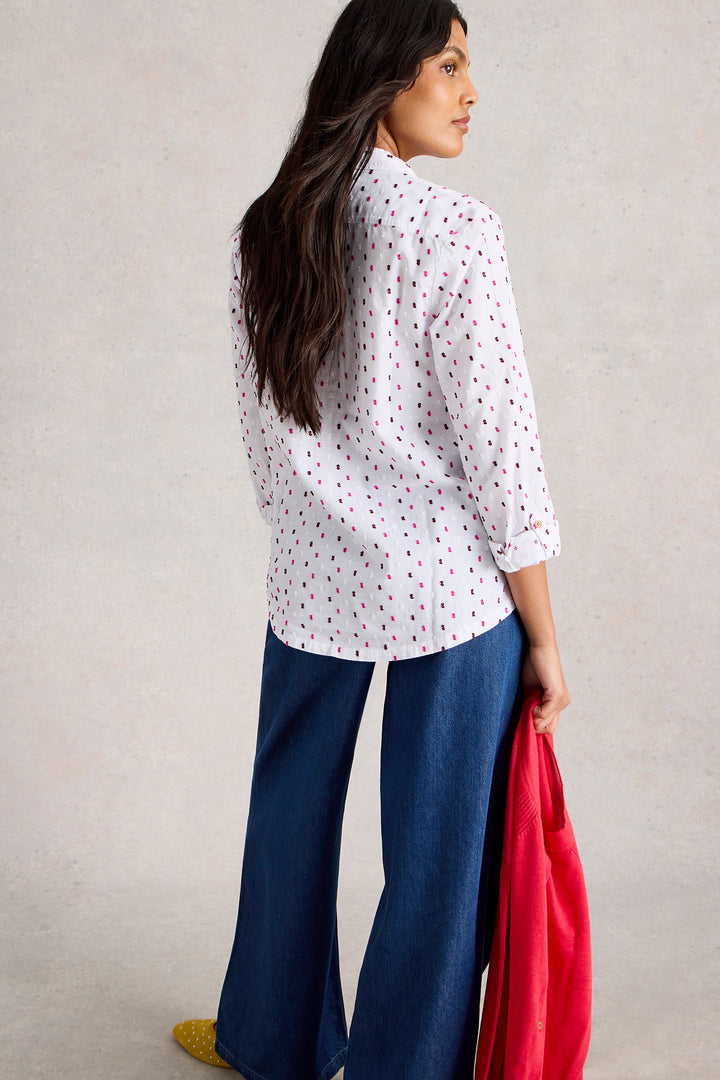 Woman wearing a white blouse with red patterns and blue pants, holding a red jacket against a neutral background.