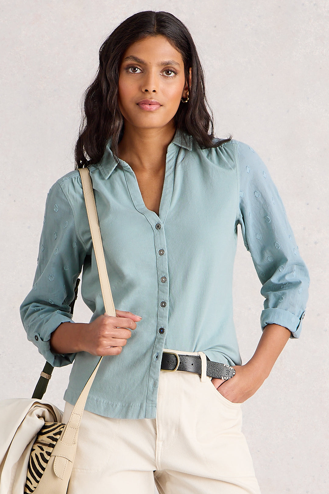 Woman wearing a light blue shirt with rolled-up sleeves, holding a beige bag against a plain background